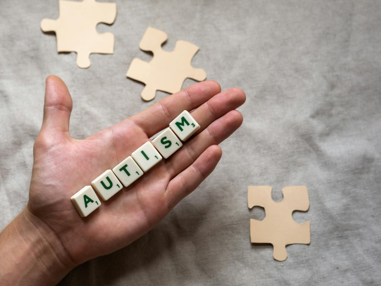 Hand displaying 'autism' spelled with scrabble tiles, accompanied by puzzle pieces, symbolizing autism awareness.